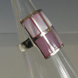 May include: A silver ring featuring a rectangular face inlaid with six vertical, rectangular pieces of iridescent pink mother-of-pearl. The inlay is arranged in two columns of three, separated by a thin silver crossbar, set within a polished silver frame.