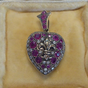 May include: A gold heart-shaped pendant with a fleur-de-lis design in the center. The pendant is adorned with red rubies and white diamonds.