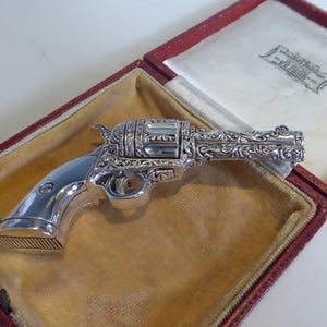Pode incluir: Um revólver prateado detalhado com ornamentos de volutas, repousando numa caixa forrada a veludo vermelho. A arma apresenta um acabamento polido e gravuras intrincadas, mostrando um design clássico. O interior da caixa é creme.