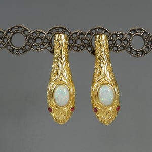 May include: A pair of gold-colored earrings with an ornate, textured design. Each earring features an oval, iridescent white stone and two small red stones. The earrings are displayed on a decorative silver-colored bar.