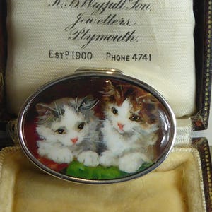 May include: A silver oval trinket box with a hinged lid. The lid features a painted image of two white kittens with brown and gray markings. The box is in a brown velvet box with a white label that reads "Est. 1900 Phone 4741".