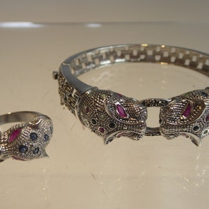 May include: A silver-coloured jewellery set comprising a ring and a bracelet. The bracelet and ring feature detailed jaguar head designs, embellished with red, blue, and green gemstones. The bracelet has a hinged clasp.