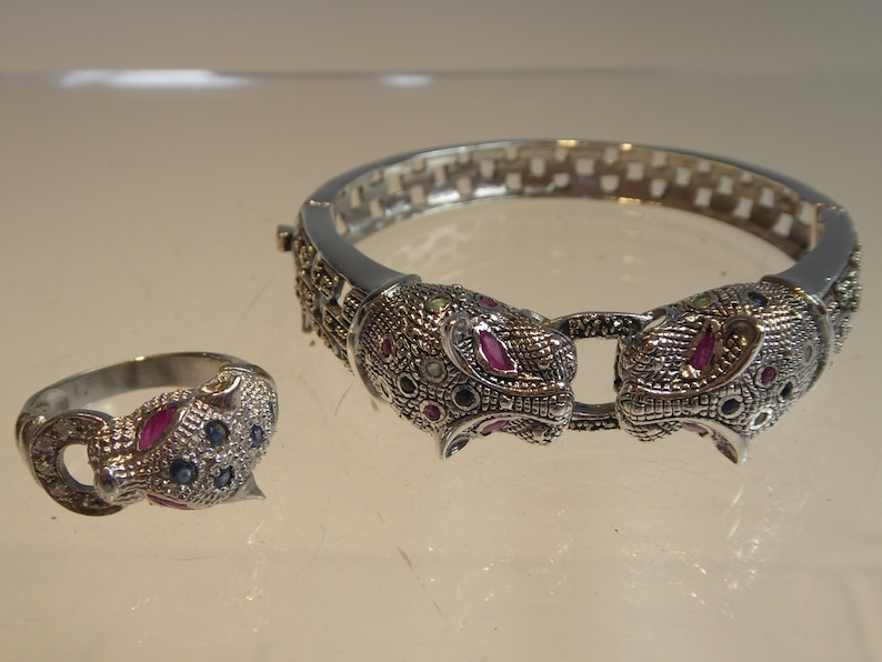 May include: A silver-coloured jewellery set comprising a ring and a bracelet. The bracelet and ring feature detailed jaguar head designs, embellished with red, blue, and green gemstones. The bracelet has a hinged clasp.
