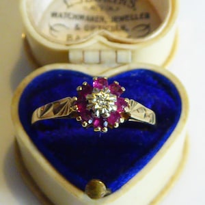 May include: A gold ring featuring a cluster of red rubies and a single diamond at its centre. The ring is displayed in a blue velvet heart-shaped box.