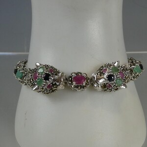 May include: A silver-toned bracelet featuring two stylized panther heads facing each other, accented with small green, red, and black stones. The links are decorated with tiny metallic beads, and a central oval red stone sits between the panther heads.