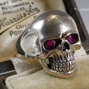 Puede incluir: Un anillo de calavera plateado con ojos color rubí, exhibido en un joyero vintage. El diseño de calavera del anillo presenta dientes y rasgos faciales detallados. La caja tiene el texto "H. Harris & Son".
