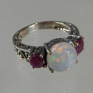 May include: Silver ring with a large, iridescent opal and two red rubies. The ring has an ornate filigree design on the band. The gemstones are set in a silver band.