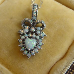 Peut inclure: Un collier pendentif en argent avec un opale en forme de cœur entourée de petites opales. Le pendentif a un design en forme de nœud et est présenté sur un fond jaune. C'est un bijou.