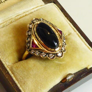 Op de afbeelding: Een gouden ring met een grote, ovaalvormige zwarte cabochonsteen in het midden, geflankeerd door twee driehoekige rode stenen. De zetting is omringd door een geschulpte rand van kleine, heldere, rondgeslepen stenen in een textuurrijke gouden zetting.