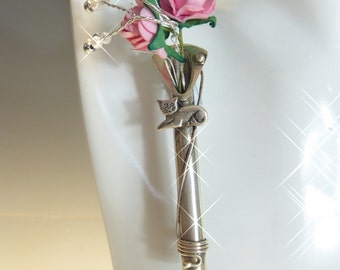 Puede incluir: Un broche de plata con un pequeño jarrón que contiene flores rosas y un dije de gato plateado. El jarrón está envuelto con alambre de plata y las flores están rodeadas de cuentas de perlas blancas.