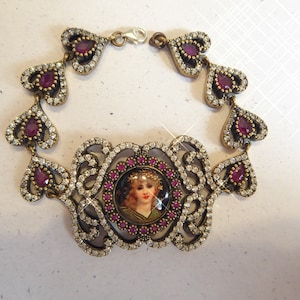 Op de afbeelding: Een zilveren armband met een hartvormig ontwerp, met een klein portret van een vrouw met een bloemenkrans. De armband is versierd met heldere en roze strass.