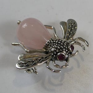 Könnte beinhalten: Eine silberne Bienenbrosche mit rosa Rosenquarz und Rubin-Akzenten. Die Flügel der Biene sind mit filigranen Designs verziert.