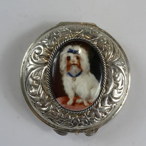 Puede incluir: Un estuche plateado con un retrato ovalado de un perro blanco con un lazo y collar azules. El estuche tiene un diseño floral ornamentado alrededor del retrato y un cierre con bisagras.