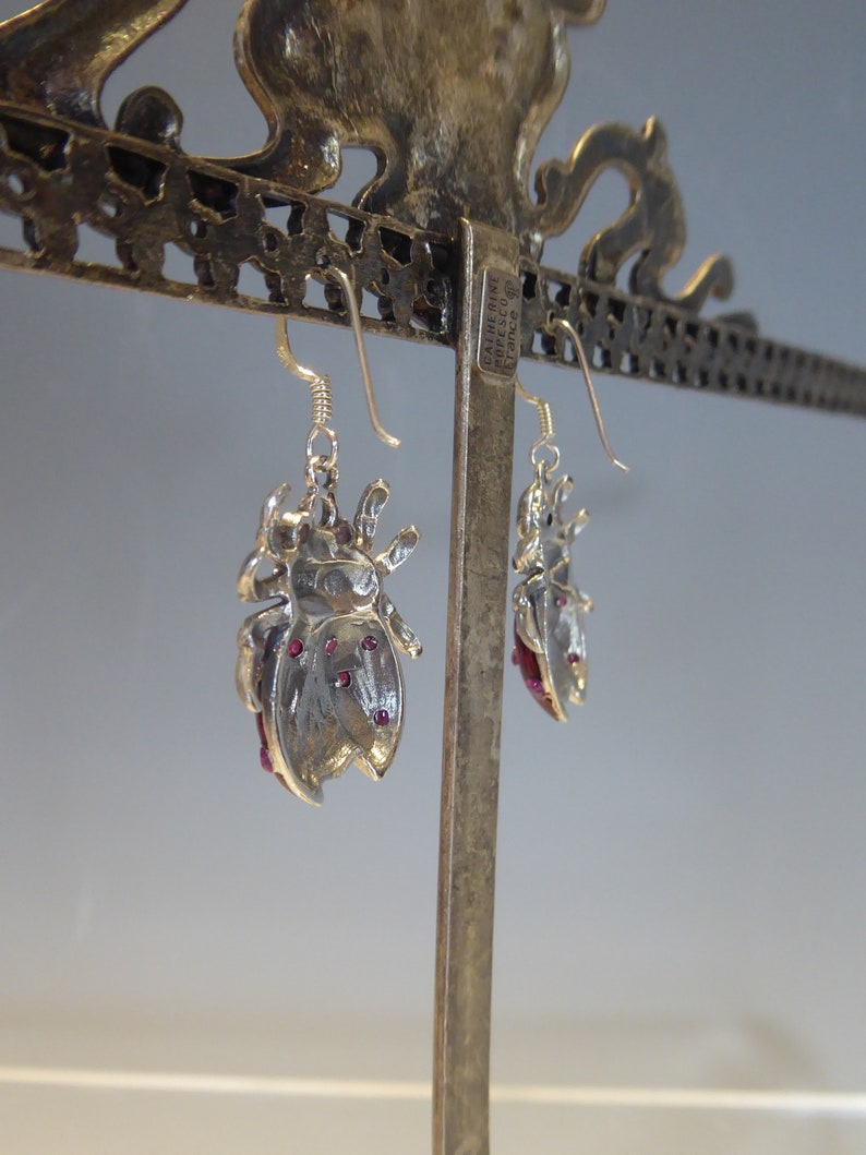 May include: Silver earrings with a detailed beetle design and pink accents. The earrings are displayed on a silver stand with the text "Catherine Appleson,  Tivoli" on the stand.