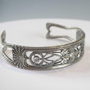 Puede incluir: Una pulsera de brazalete de metal en tono plateado con un intrincado diseño calado, patrones en relieve en forma de abanico y pequeños recortes circulares. La pulsera está acentuada con varios diamantes de imitación redondos, claros y brillantes.