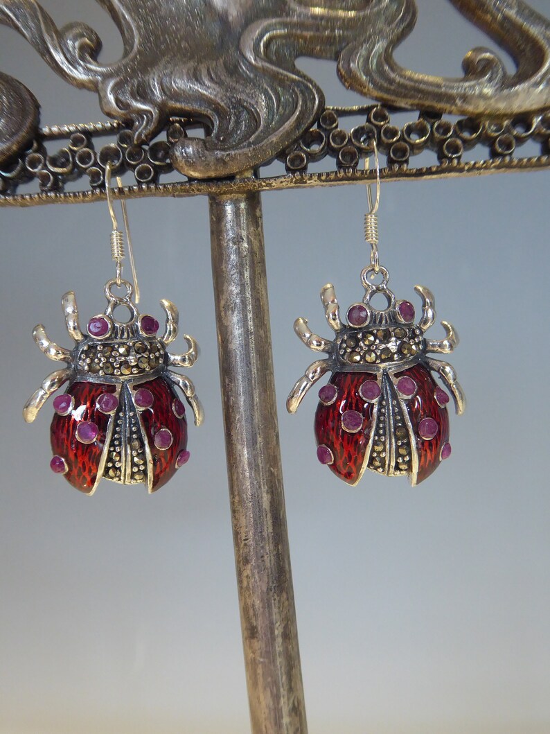 May include: A pair of silver earrings with a ladybug design. The ladybugs are red with black spots and have pink gemstones for eyes. The earrings are made with marcasite stones.