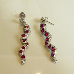 May include: A pair of silver-toned drop earrings in a serpentine shape, each featuring a series of round, vibrant red stones set along the length of the body, accented by small, clear pavé-set stones throughout the entire curved metal surface.