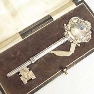 May include: A silver key with ornate details, including a circular medallion with an inscription, is displayed in a black velvet-lined box.
