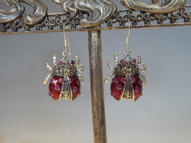 May include: A pair of silver earrings shaped like ladybugs with red enamel and purple gemstone accents.