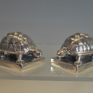 Puede incluir: Dos figuritas de tortuga de plata sobre bases cuadradas. Las tortugas se miran y tienen patrones de caparazón detallados.