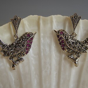 May include: A pair of silver bird earrings with a detailed design and purple gemstones.