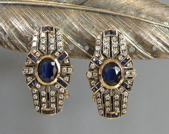 May include: A pair of gold earrings with a geometric design featuring a large blue sapphire surrounded by smaller diamonds and sapphires.