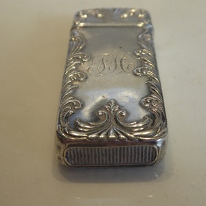 Puede incluir: Un porta-cerillas de plata con un diseño ornamentado y un monograma grabado. El porta-cerillas tiene una tapa con bisagras y un fondo acanalado.