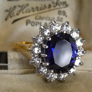 May include: A gold ring with a large, oval, deep blue sapphire encircled by smaller, clear gemstones. The ring is set against a beige background, with the text "H. Harris & Son" visible.