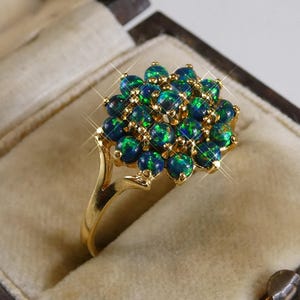 Könnte beinhalten: Ein goldener Ring mit einem Cluster aus runden, irisierenden Edelsteinen in Grün- und Blautönen. Der Ring ist in einem goldenen Band gefasst und wird in einer Schmuckschatulle präsentiert.