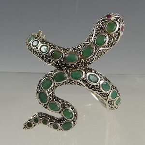 May include: A silver-toned snake ring with green and red gemstones. The snake's body is adorned with oval-shaped green stones, and the head features two red stones. The ring has a detailed, textured design.