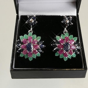 Puede incluir: Un par de pendientes colgantes de tono plateado con un diseño de racimo floral. Cada pendiente muestra un zafiro azul ovalado central rodeado por pétalos de rubíes rosas y esmeraldas verdes ovaladas, con un pequeño broche de flor.