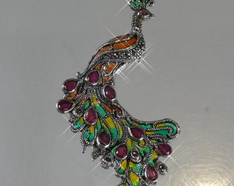 Puede incluir: Broche de pavo real plateado con un diseño detallado. El broche presenta un diseño de esmalte colorido en naranja, verde y amarillo, acentuado con piedras preciosas rojas. La corona del pavo real es verde y las plumas de la cola están desplegadas.