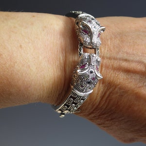 May include: Silver bracelet with two panther head motifs, adorned with red and black gemstones. The bracelet, with its intricate design, is shown on a wrist. This is a piece of jewellery.