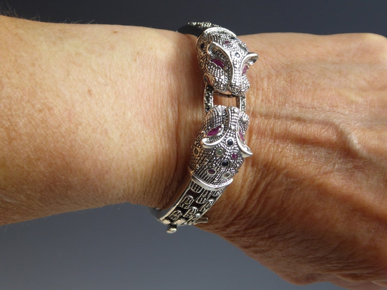 May include: Silver bracelet with two panther head motifs, adorned with red and black gemstones. The bracelet, with its intricate design, is shown on a wrist. This is a piece of jewellery.