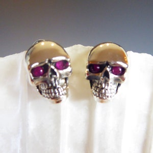 Könnte beinhalten: Ein Paar silberne Totenkopf-Ohrstecker mit rosafarbenen Edelsteinaugen.