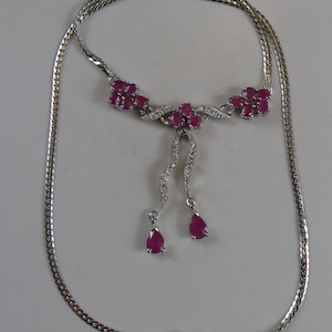May include: A silver-toned necklace featuring a central cluster of oval-cut deep pink rubies and small clear stones. Two curved, stone-encrusted stems extend downward, each ending in a single pear-shaped ruby drop, attached to a flat chain.