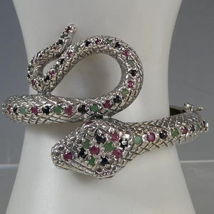 Könnte beinhalten: Ein silbernes Schlangenarmband mit strukturierter Oberfläche. Kopf und Schwanz der Schlange winden sich, verziert mit farbigen Edelsteinen in Rot, Grün und Schwarz. Das Armband wird auf einer weißen Oberfläche präsentiert.