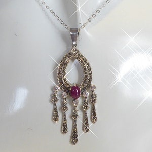Peut inclure: Un collier en argent avec un pendentif qui pend et qui présente une pierre précieuse rouge entourée de pierres claires et un motif détaillé de marcassite.