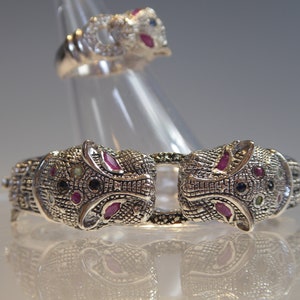 May include: A silver-coloured bracelet and ring set. The bracelet features two animal head designs with red, blue, and black gemstone accents. The matching ring has a similar design. The jewellery is displayed on a clear stand.