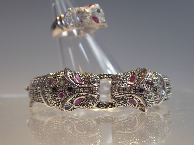 May include: A silver-coloured bracelet and ring set. The bracelet features two animal head designs with red, blue, and black gemstone accents. The matching ring has a similar design. The jewellery is displayed on a clear stand.