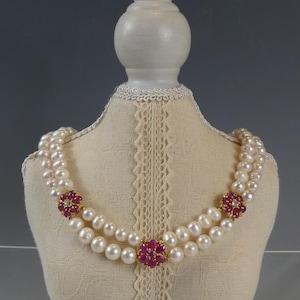 Pode incluir: Um colar de pérolas de duas voltas com três detalhes florais de rubis e diamantes. O colar é exibido em um busto de manequim creme com uma borda de renda.