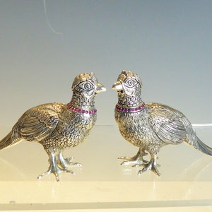 Può includere: Due figurine di fagiano d'argento con accenti rosso rubino. I fagiani sono dettagliati con piume testurizzate e hanno code lunghe.