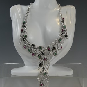 May include: A silver necklace with a peacock feather design. The necklace is adorned with green and red gemstones and black marcasite.