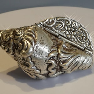 Puede incluir: Una caja de concha de mar plateada, con bisagras, con intrincados detalles. La caja está decorada con motivos florales y tiene una tapa con bisagras.