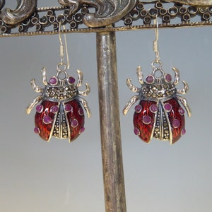 May include: A pair of silver earrings shaped like ladybugs with red enamel and purple gemstones. The earrings are hanging from a silver stand.