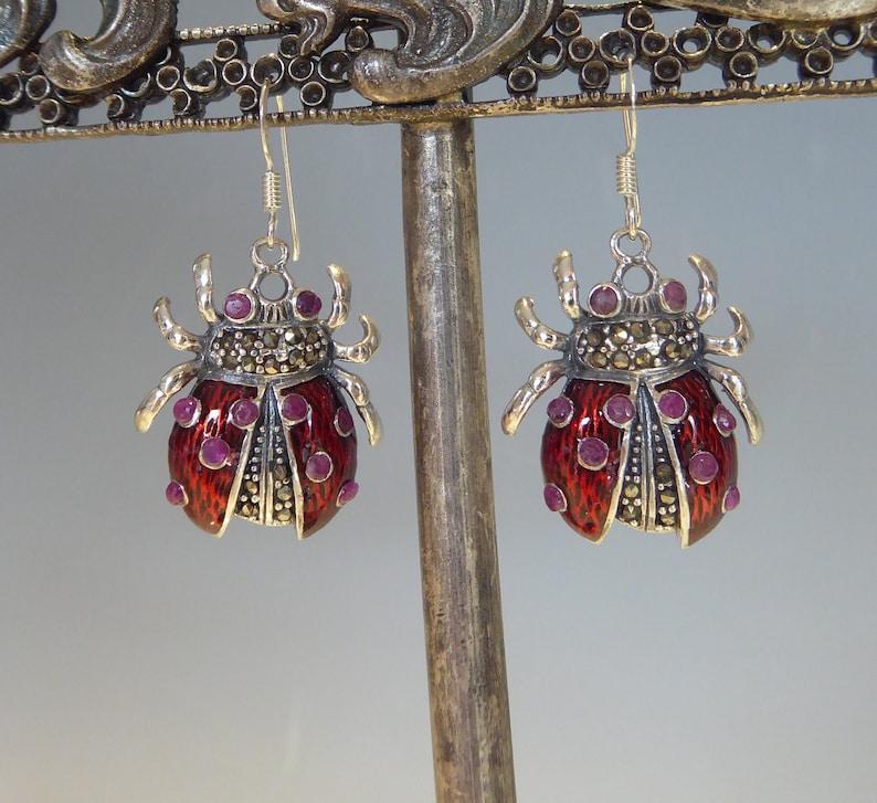 May include: A pair of silver earrings shaped like ladybugs with red enamel and purple gemstones. The earrings are hanging from a silver stand.
