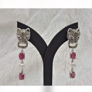 May include: A pair of silver dangle earrings featuring a black panther head design. The panther head is encrusted with small black stones and has red eyes. The earrings dangle with three red stones and a white pearl.