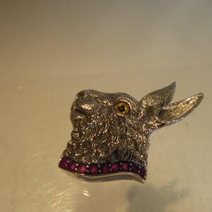 May include: Silver rabbit brooch with a textured fur design and a single yellow eye. The rabbit is wearing a collar of pink gemstones.