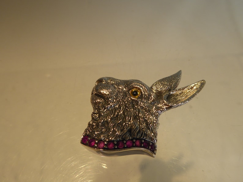 May include: Silver rabbit brooch with a textured fur design and a single yellow eye. The rabbit is wearing a collar of pink gemstones.
