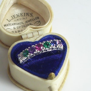 Puede incluir: Un anillo de plata con una fila de piedras preciosas de colores, presentado en un joyero en forma de corazón con interior de terciopelo azul. La caja crema tiene el texto "L. Jenkins, Watchmaker, Jeweller & Optician, ARC".